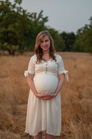 A maternity portrait in a golden field features a woman in a white dress at sunset.