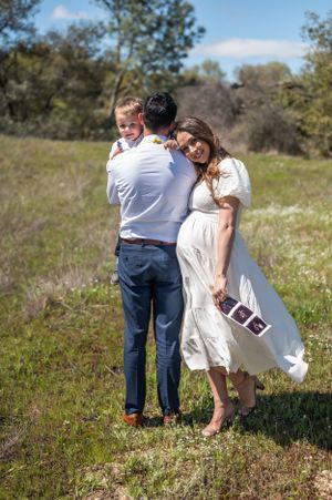 A couple embraces in a sunny field while holding ultrasound photos in a sweet pregnancy announcement moment.