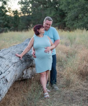 A romantic maternity photo session features a couple in light blue outfits posing by a fallen log at sunset.