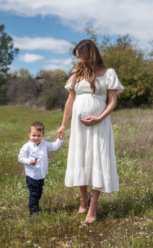 Mother in white dress holds hands with small child in flowery field.