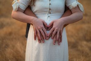 Closeup of hands forming a heart shape over a pregnant belly in a white dress.