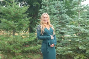 A person in a teal dress poses in front of evergreen trees during a maternity photoshoot.