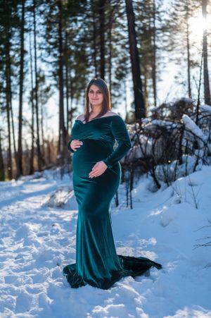 A stunning maternity photo featuring an emerald velvet gown against a snowy winter forest backdrop.