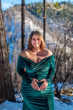 Woman wearing green velvet gown makes heart shape with hands against snowy mountain backdrop.