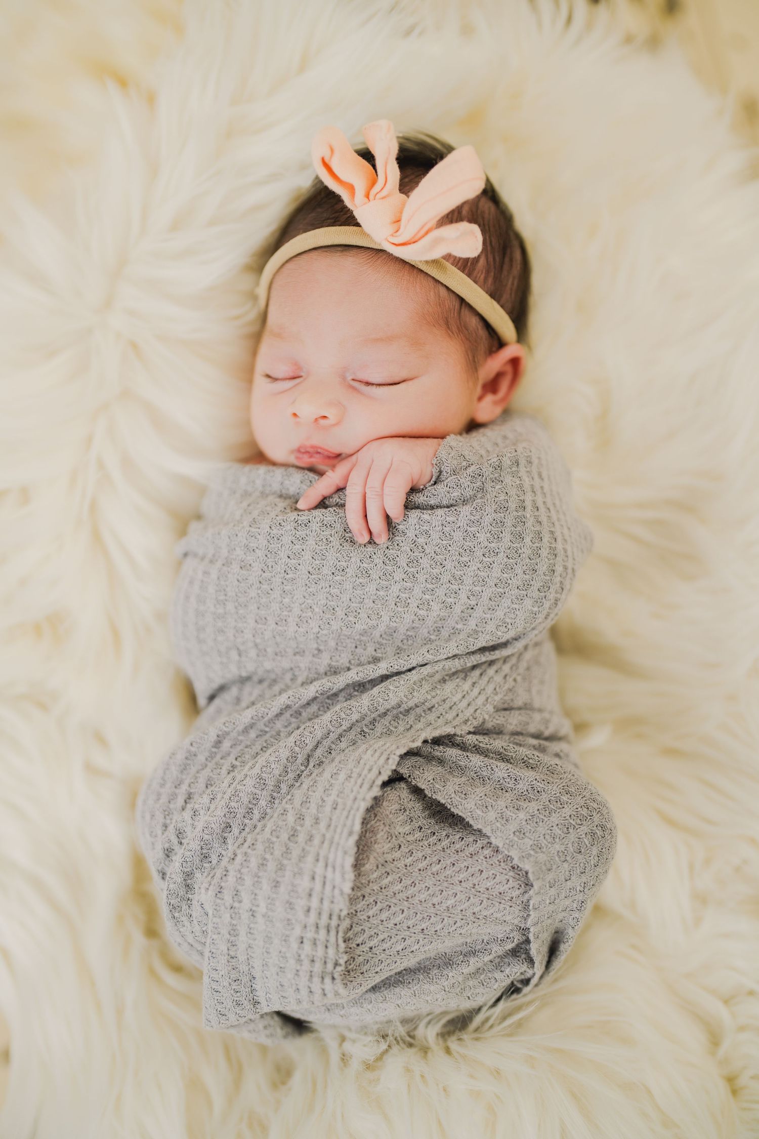 Sleeping newborn wrapped in gray knit blanket wearing pink bow headband on white fur.