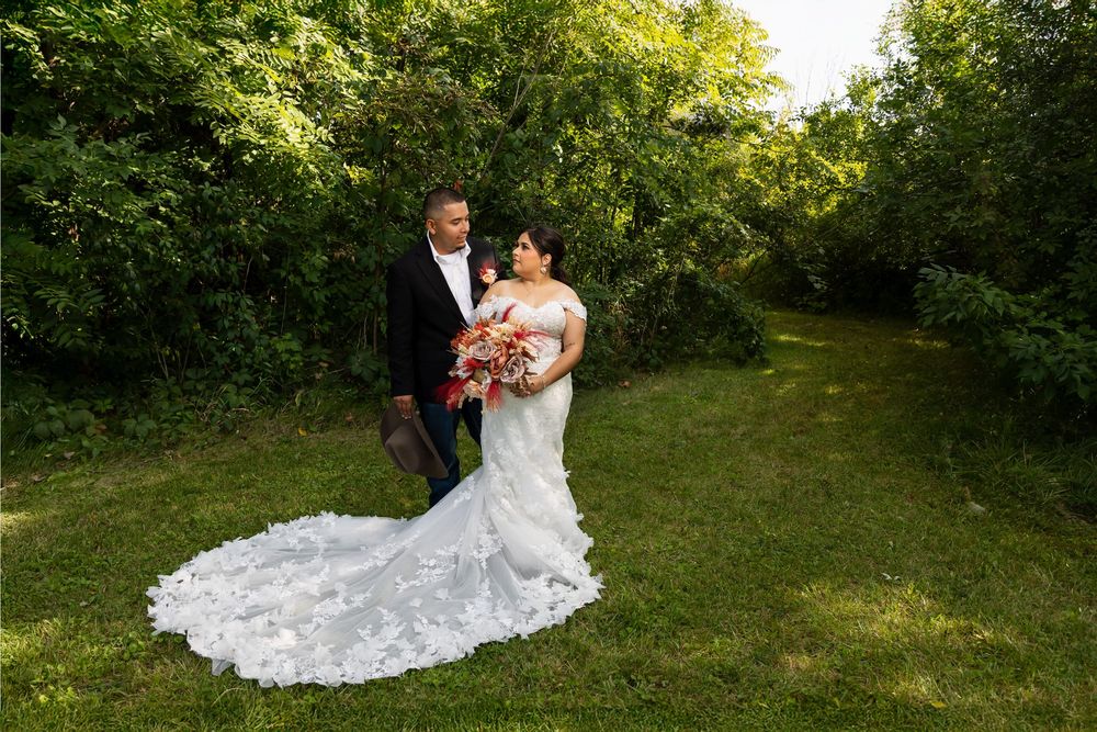 Romantic wedding portrait of a bride and groom standing in a lush green forest. The bride's intricate lace wedding dress train is spread acr