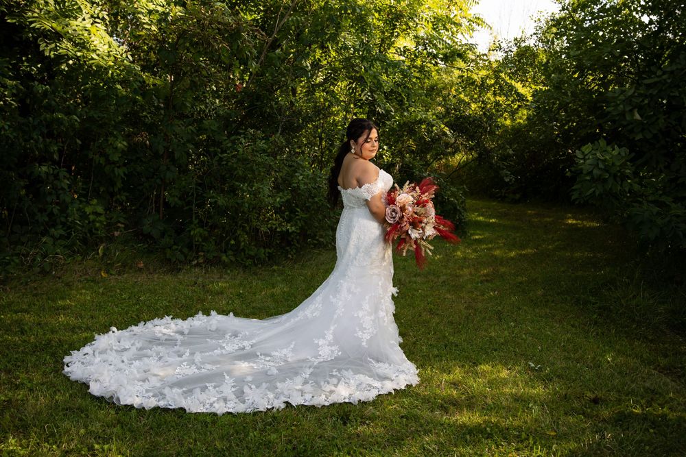 A bride in a stunning white lace gown with a long train, standing in a lush green field during golden hour. Professional bridal portrait by