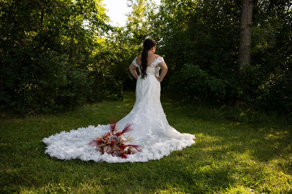 Wide shot of a bride from behind, showcasing a long, intricate lace wedding dress train spread out on green grass. Beautiful outdoor bridal