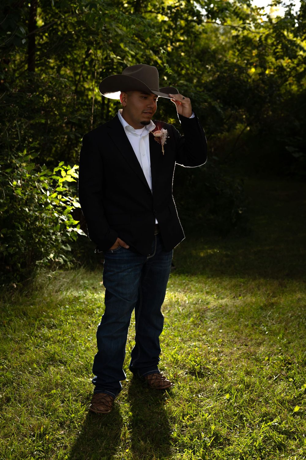 Full-body portrait of a groom wearing a cowboy hat and suit, standing in a sun-drenched forest setting for a rustic outdoor wedding. Capture