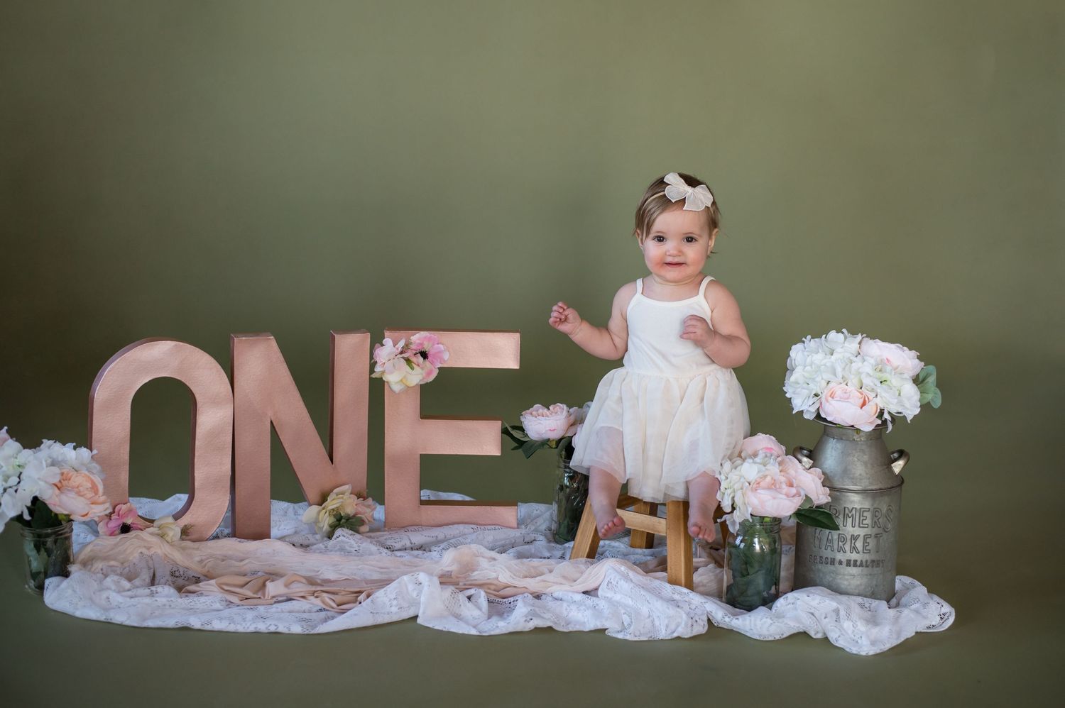 Baby in white dress standing near rose gold 'ONE' letters and floral decorations.