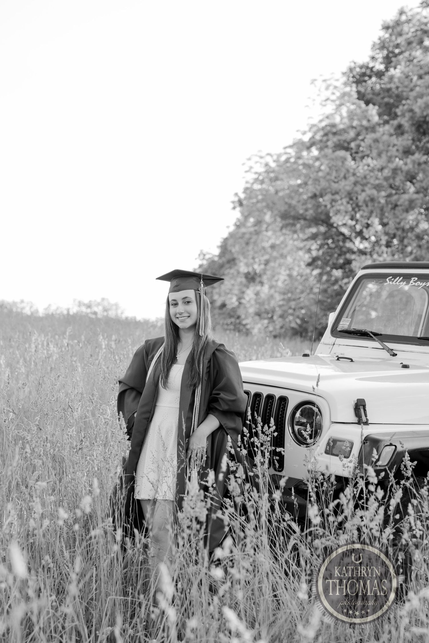 Black and white photo of graduate in cap and gown standing by vintage Jeep in meadow.