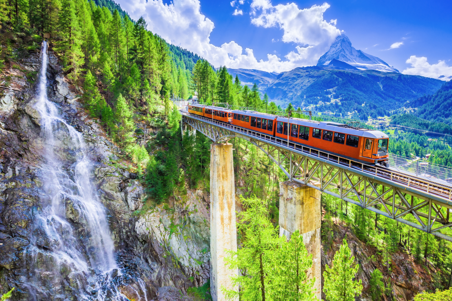 Red mountain train crosses tall concrete bridge next to cascading waterfall in Swiss Alps beneath snowy peak.