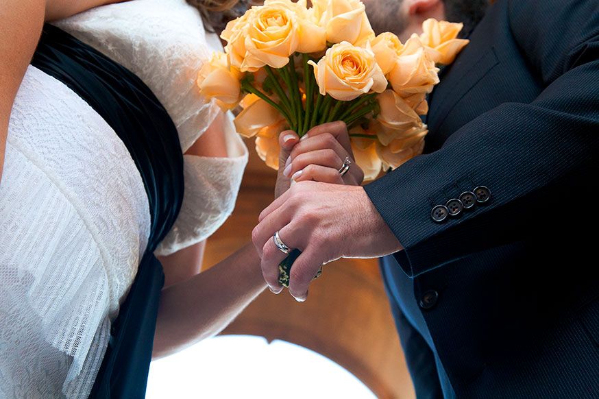 Close-up detail of wedding couple's hands with peachy rose bouquet.