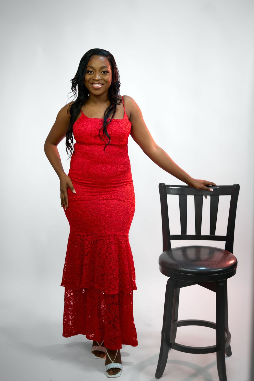 Studio portraits in red lace dress posing with black chair against white background.