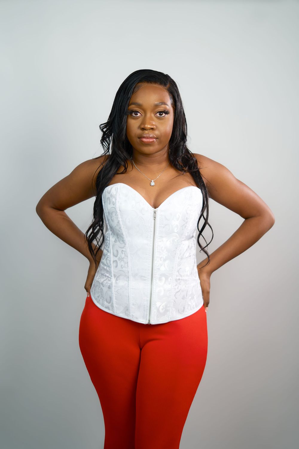 Model in white corset top and red pants poses confidently against light grey backdrop.