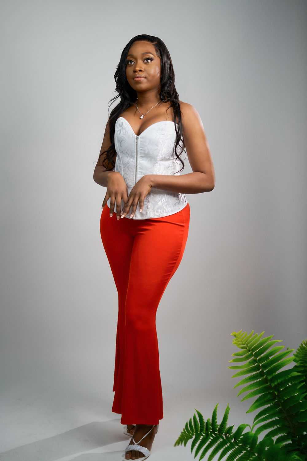 Fashion model wearing white lace corset and red flared pants standing next to decorative fern.