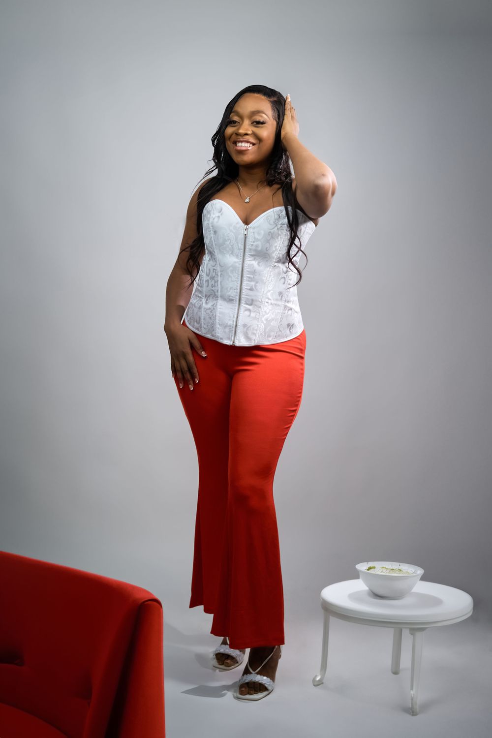 Stylish portrait featuring white corset top, red pants and casual pose near small side table.