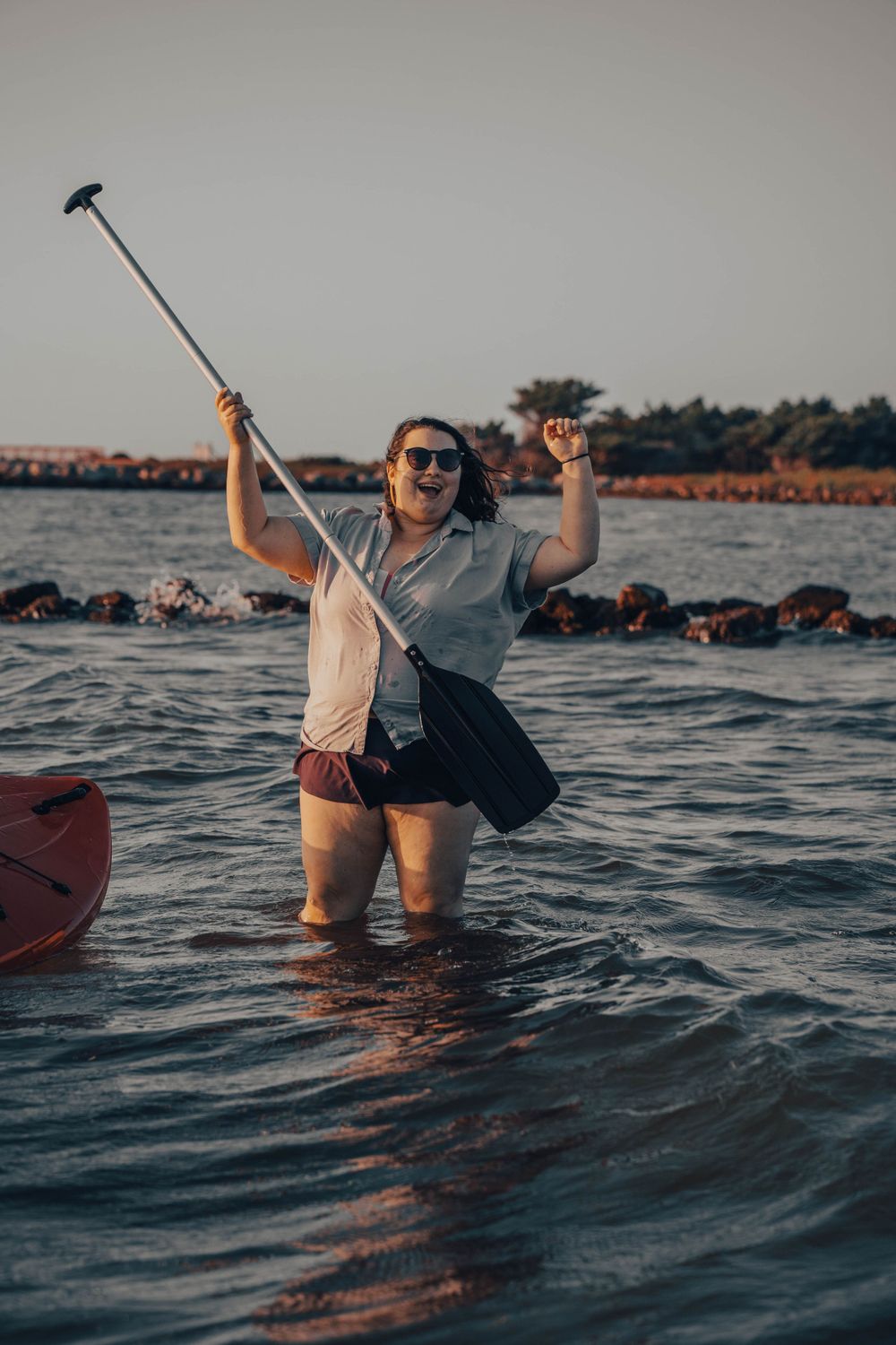 Person stands in water holding selfie stick while celebrating near shoreline at sunset.