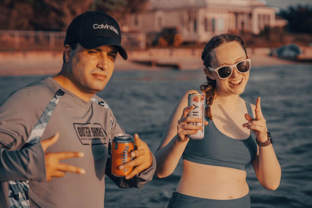 Two people holding drinks pose together on beach at sunset.