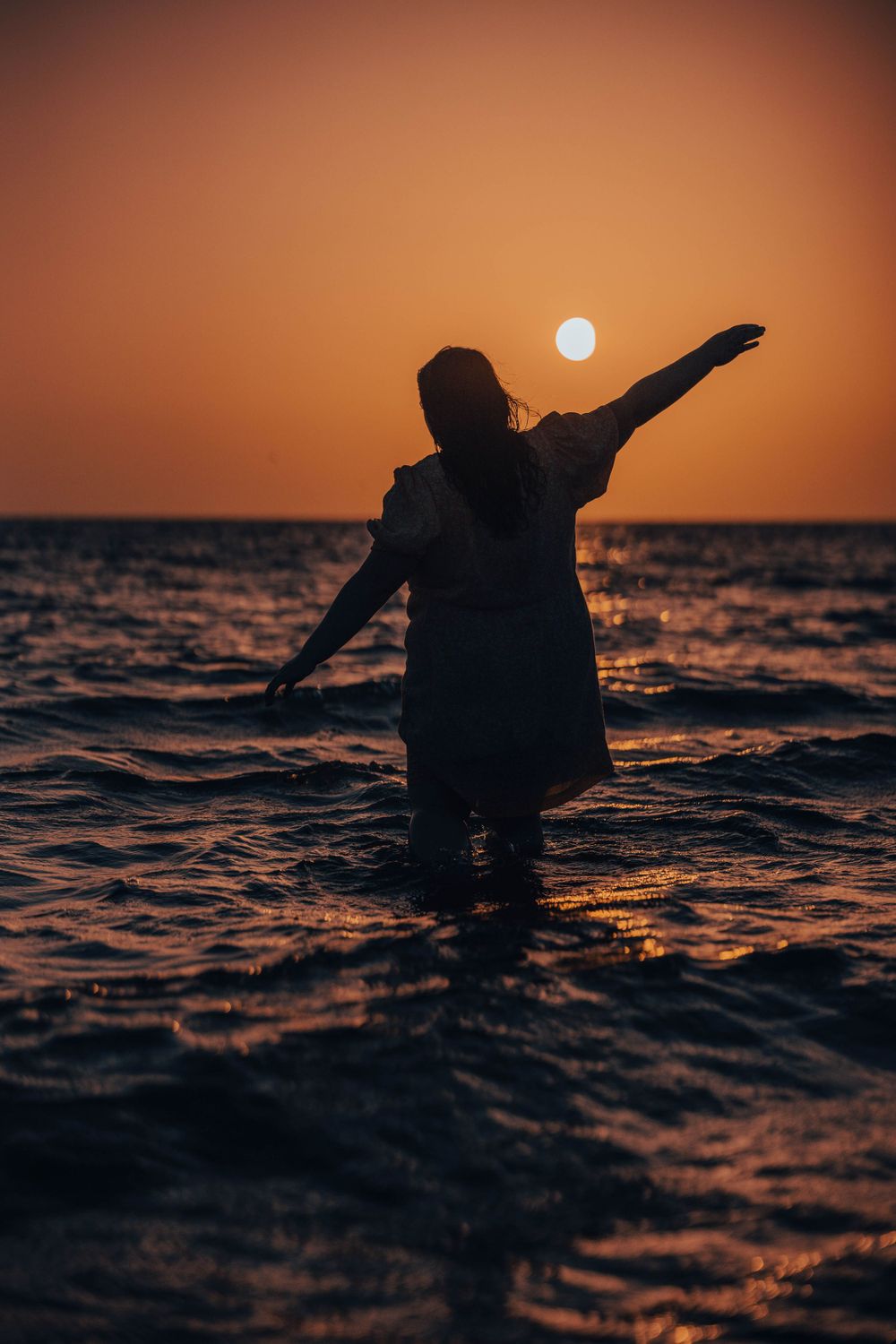 Silhouette of person with outstretched arms standing in ocean at sunset.