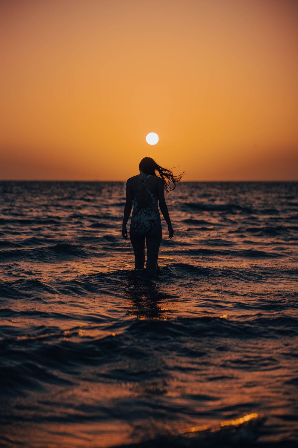 Silhouetted figure stands in ocean waves during golden sunset hour.