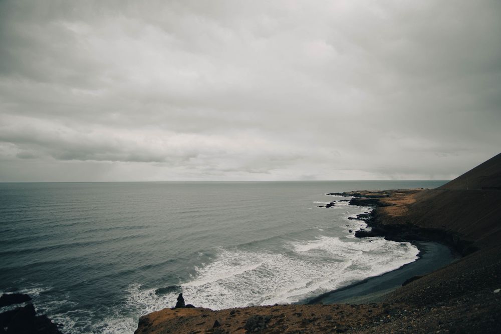 Moody coastal landscape with crashing waves against rocky cliffs under overcast skies.