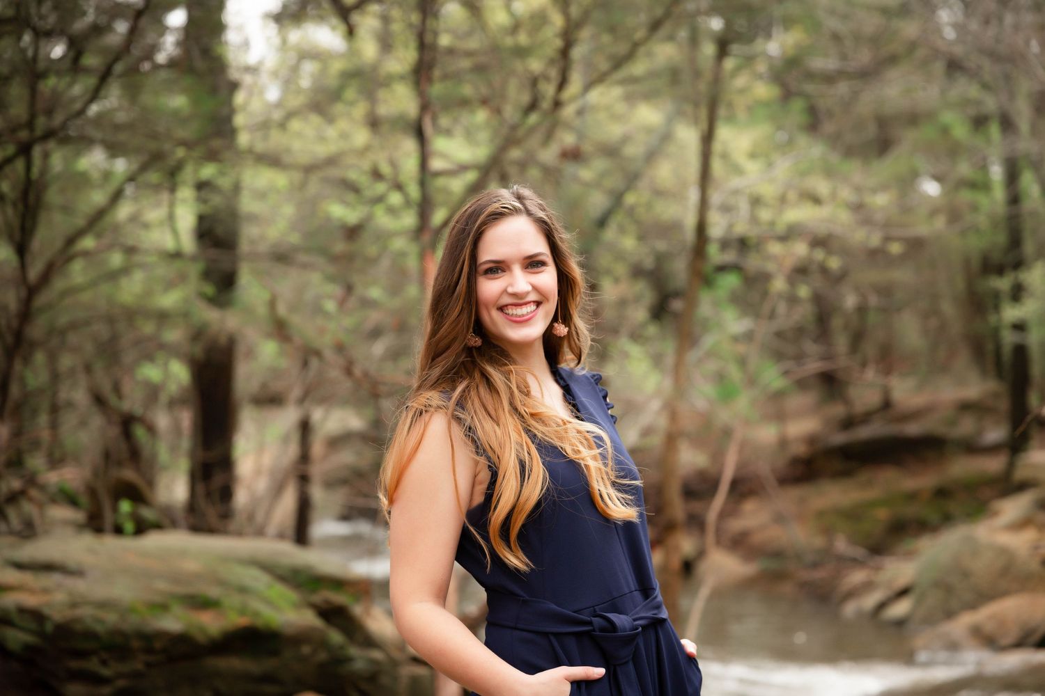 Senior girl in navy dress standing outdoors near wooded stream area.