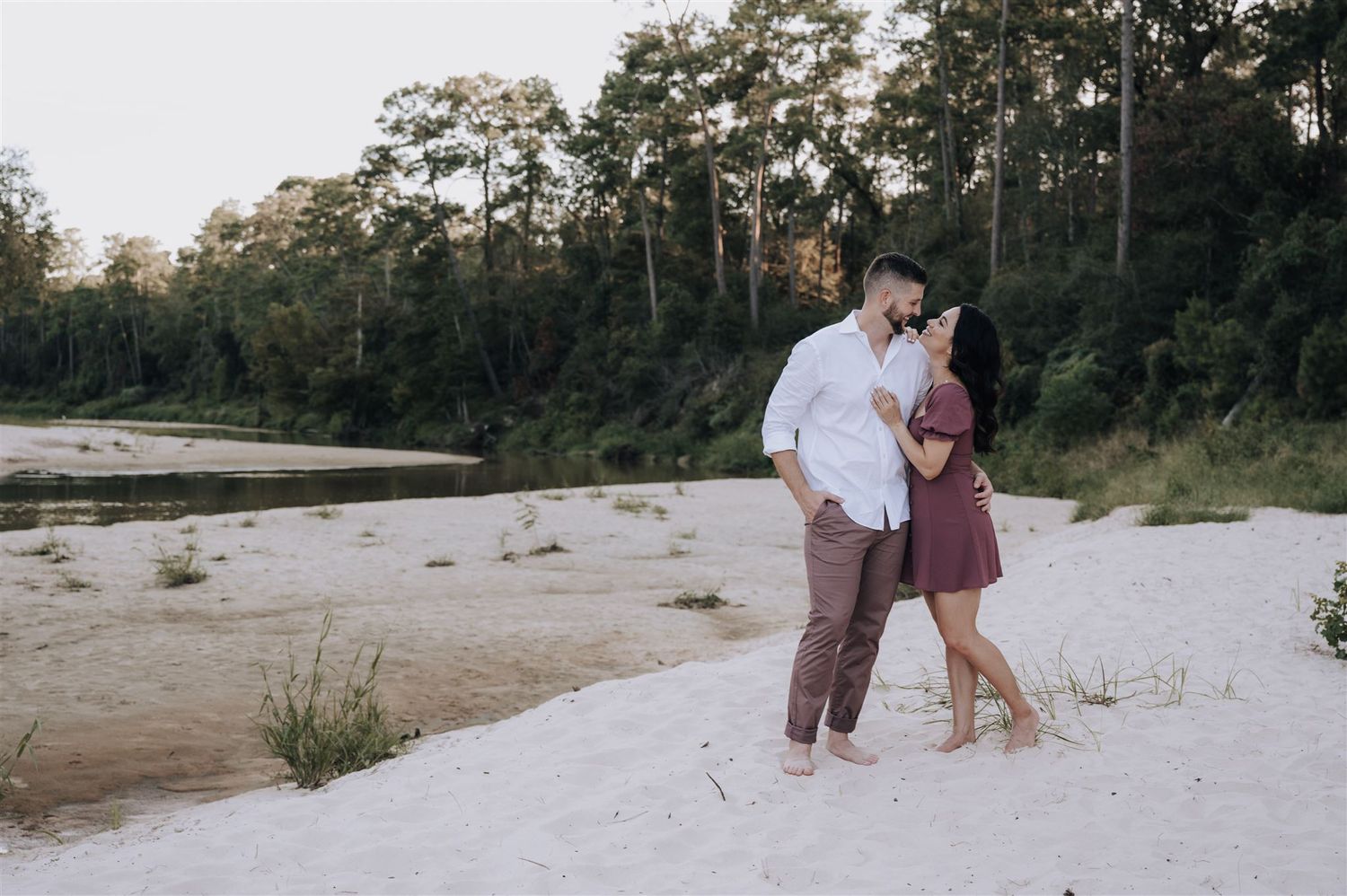 Romantic couple embracing on a sandy riverbank surrounded by trees at sunset.