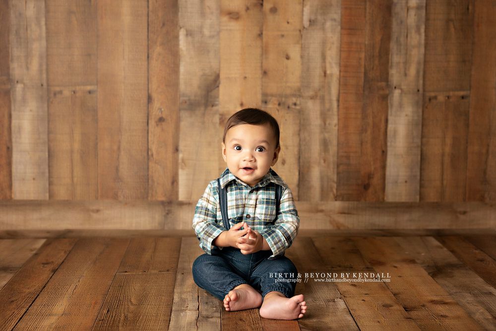 Studio portrait of a small child in plaid shirt and jeans sitting on rustic wooden floor. Kendall Kuss Photography