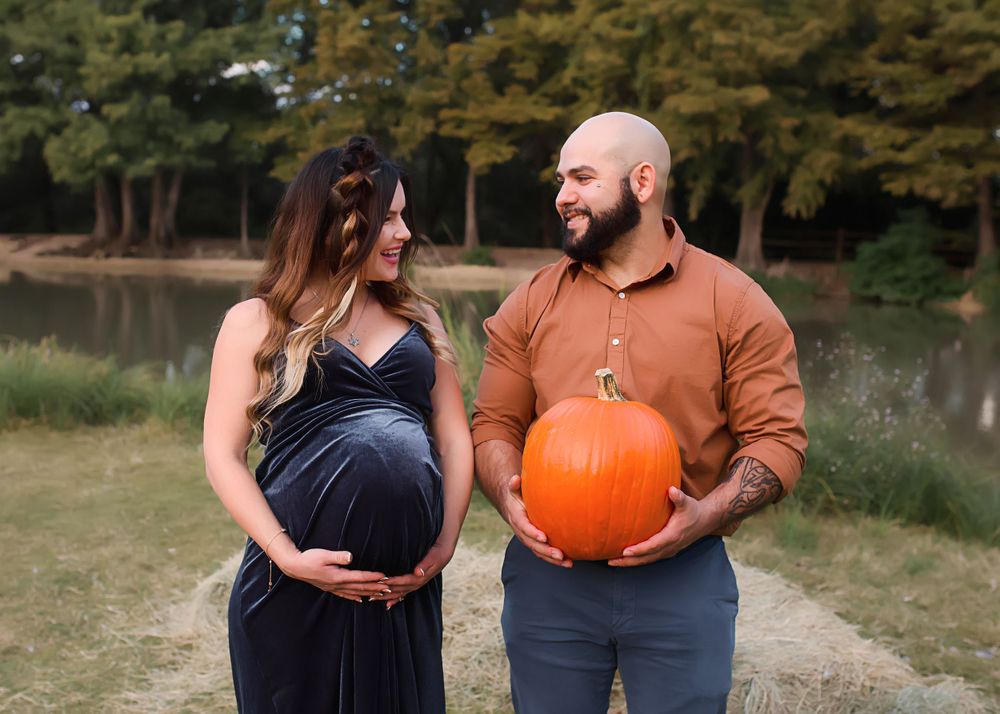 Couple in a park setting holding a large orange pumpkin during maternity photoshoot.