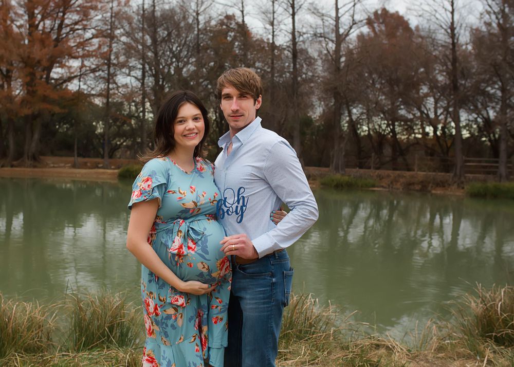 Maternity couple poses by pond, woman wearing floral blue dress and man in white shirt and jeans.