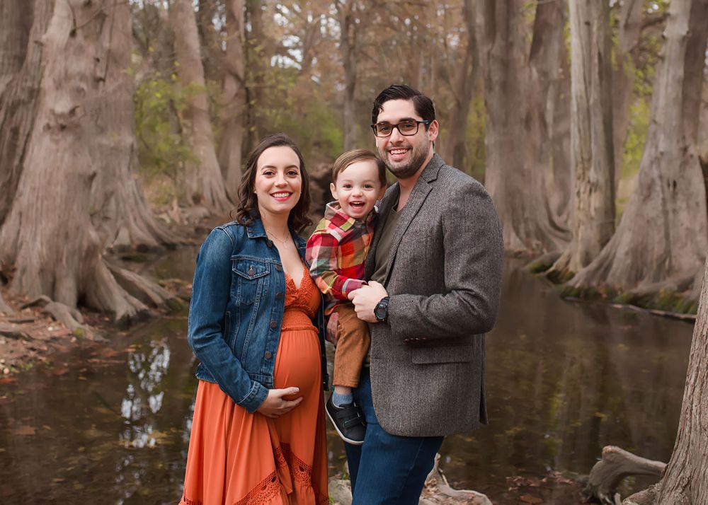 Family poses together by cypress trees near water during autumn maternity session.