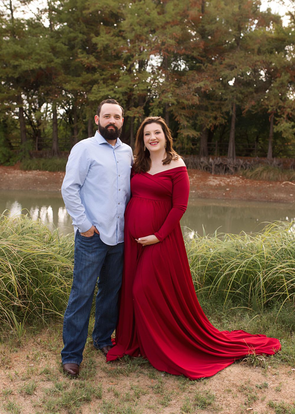 Romantic maternity photo by pond with flowing red gown and natural autumn backdrop.