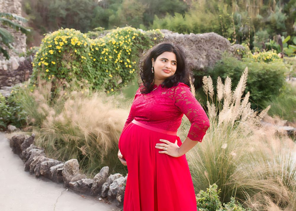 Maternity portrait of woman in red dress standing among ornamental grasses and yellow flowers.