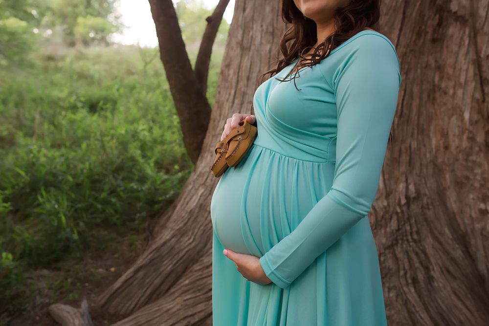 Side view of pregnant woman in turquoise dress against tree trunk.