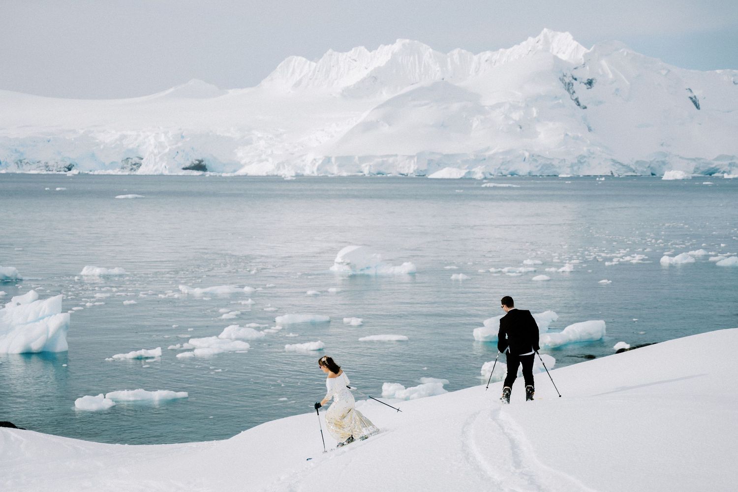 Two figures walking across snowy terrain with icy waters and glaciers in the distance.