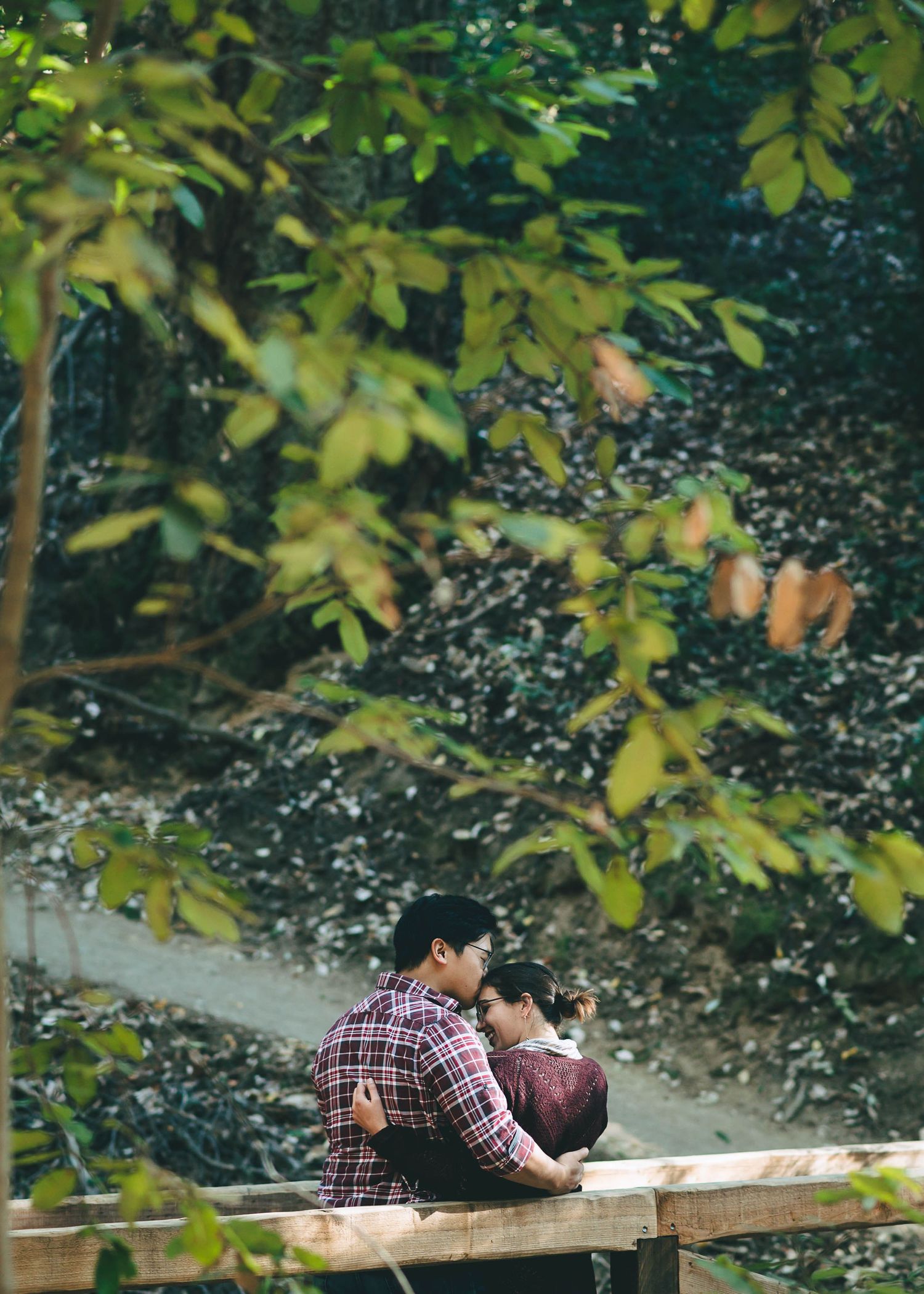 Couple sitting close together on wooden bench surrounded by green foliage in a park.