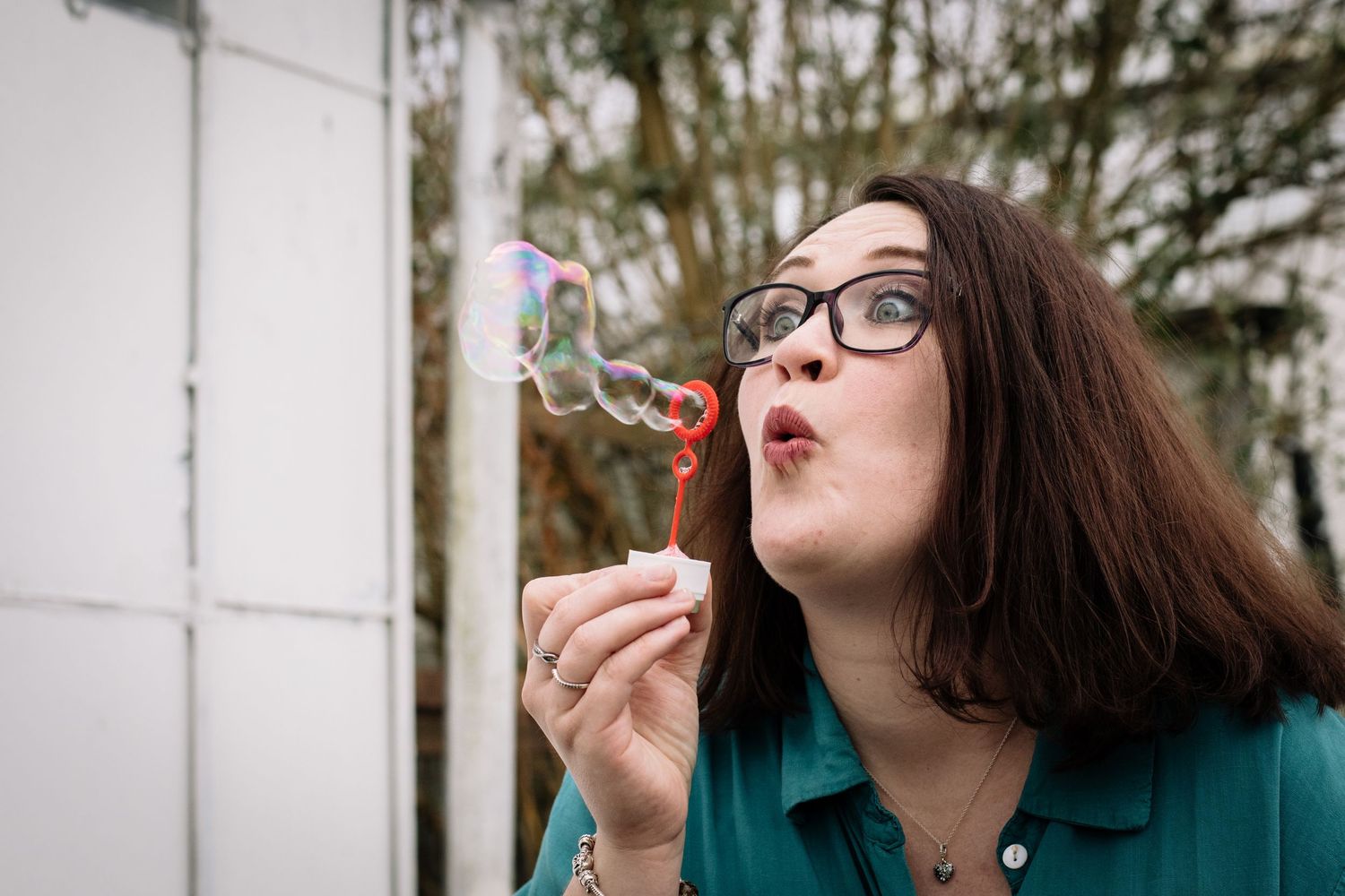 Woman in teal blazer and glasses blowing soap bubbles against a white wall.