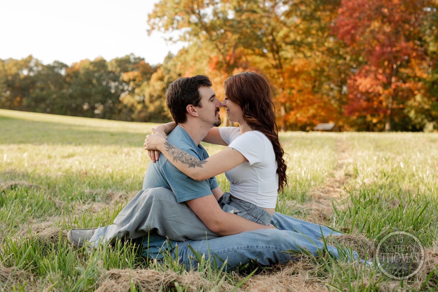 A couple embraces on a grassy hill with autumn foliage in the background.