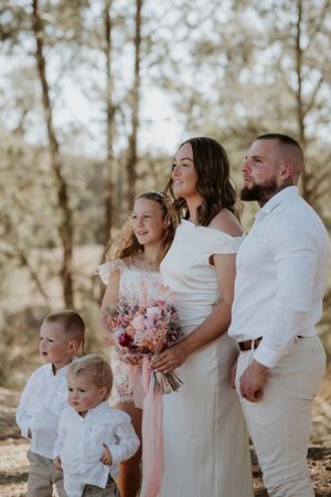 Family in white outfits pose together outdoors with pink floral bouquet in natural setting.