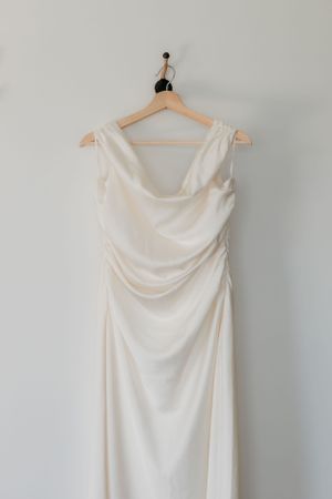 An elegant white wedding dress hangs on a wooden hanger against a light gray wall.