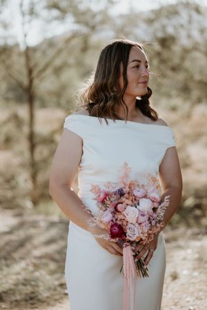 A bride in a sleeveless white gown holds a pink and burgundy bouquet in natural light.