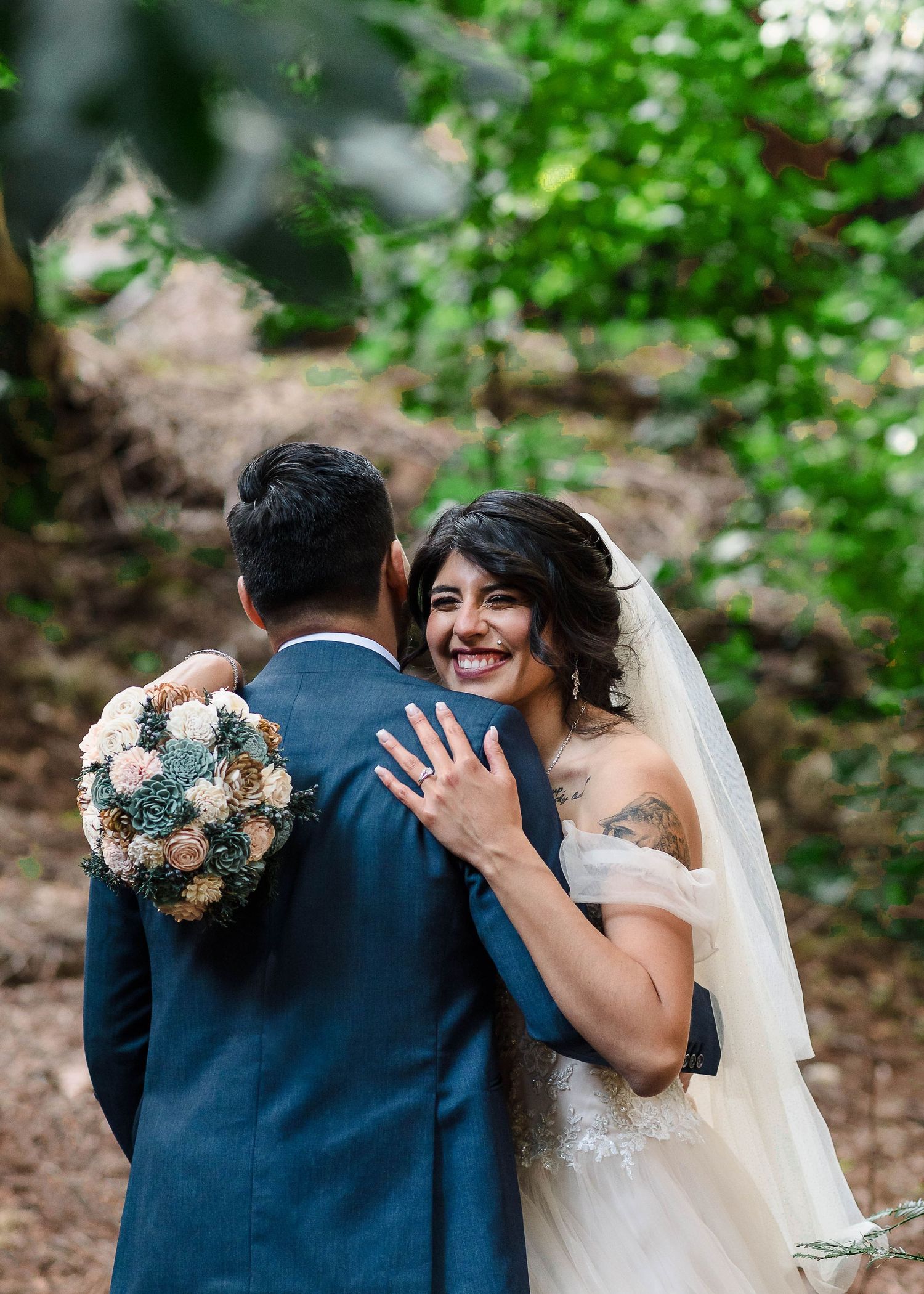 A bride in a flowing white dress shows off her ring while embracing on a forest path.