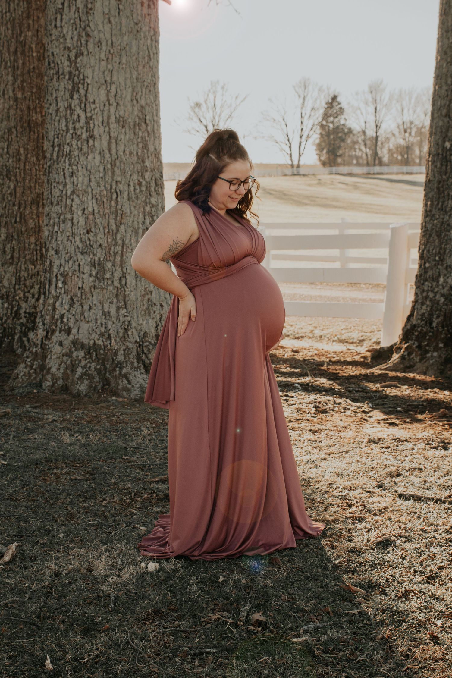 Woman in mauve dress sitting thoughtfully against large tree trunk.