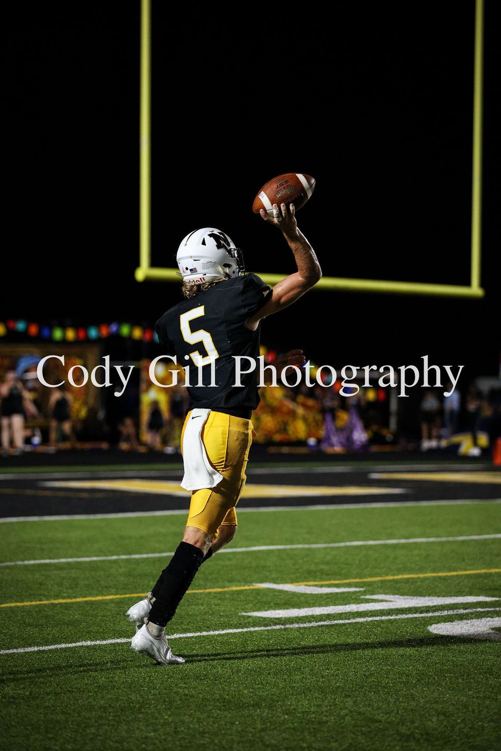 Football player in yellow pants and white jersey leaps to catch the ball near goal posts at night.