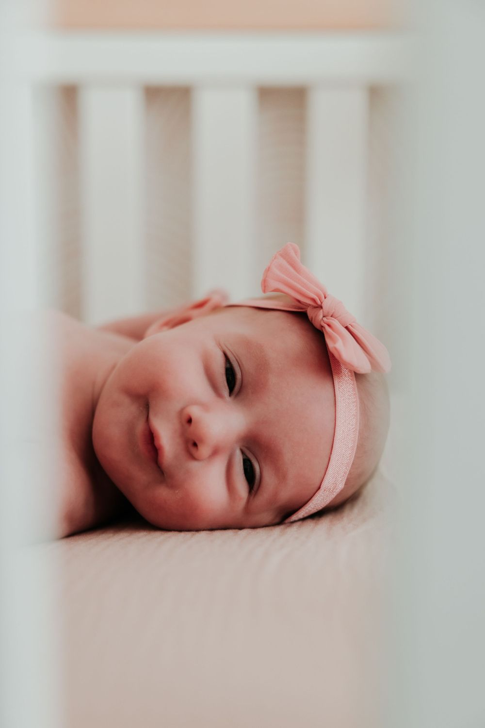 Newborn baby wearing pink bow headband lying in crib with sweet expression.