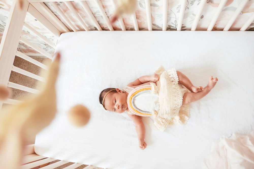 Baby in white onesie lies peacefully in a white wooden crib with natural light streaming in.