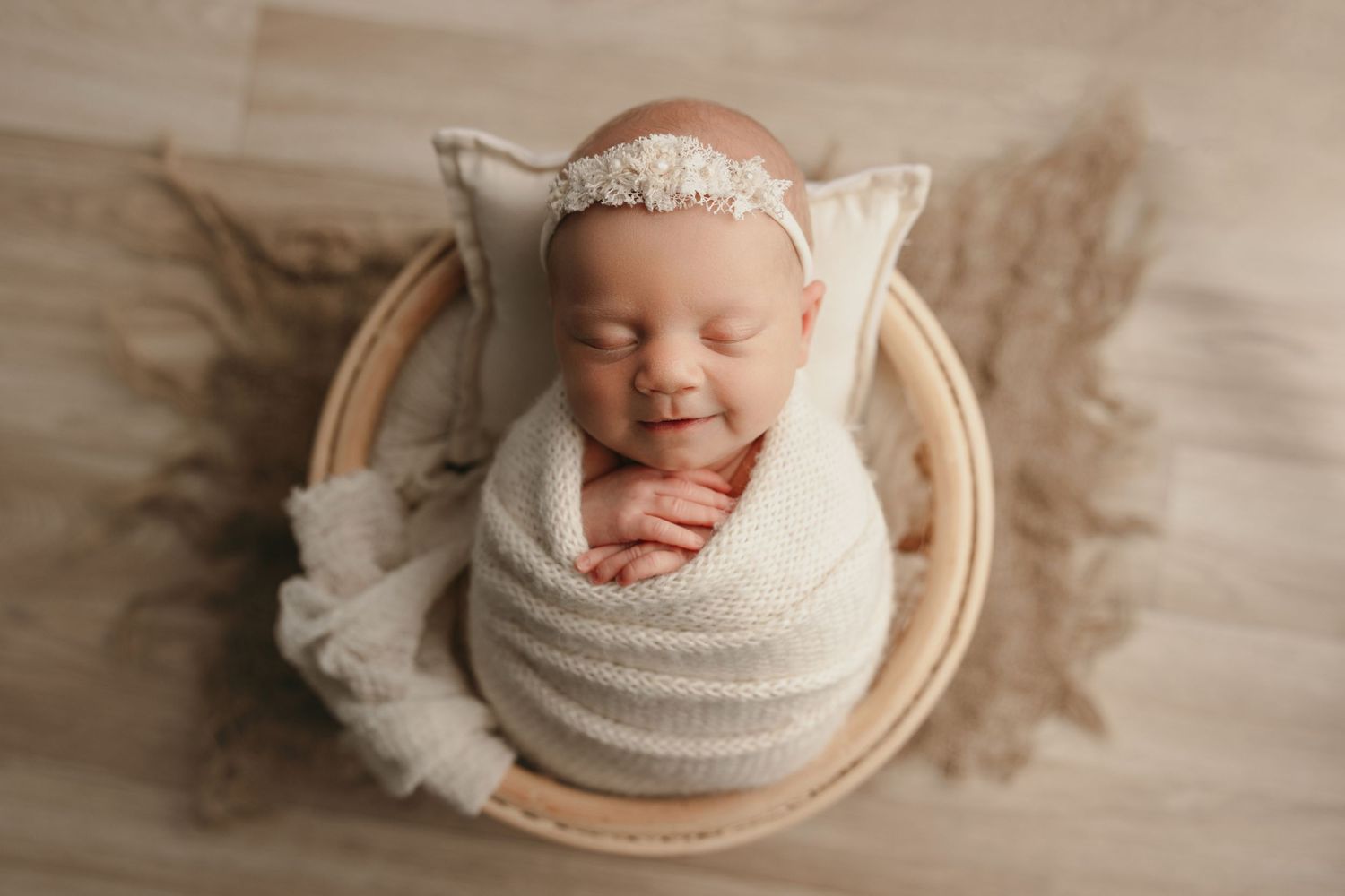 Sleeping newborn wrapped in white with lace headband in round wooden bowl.