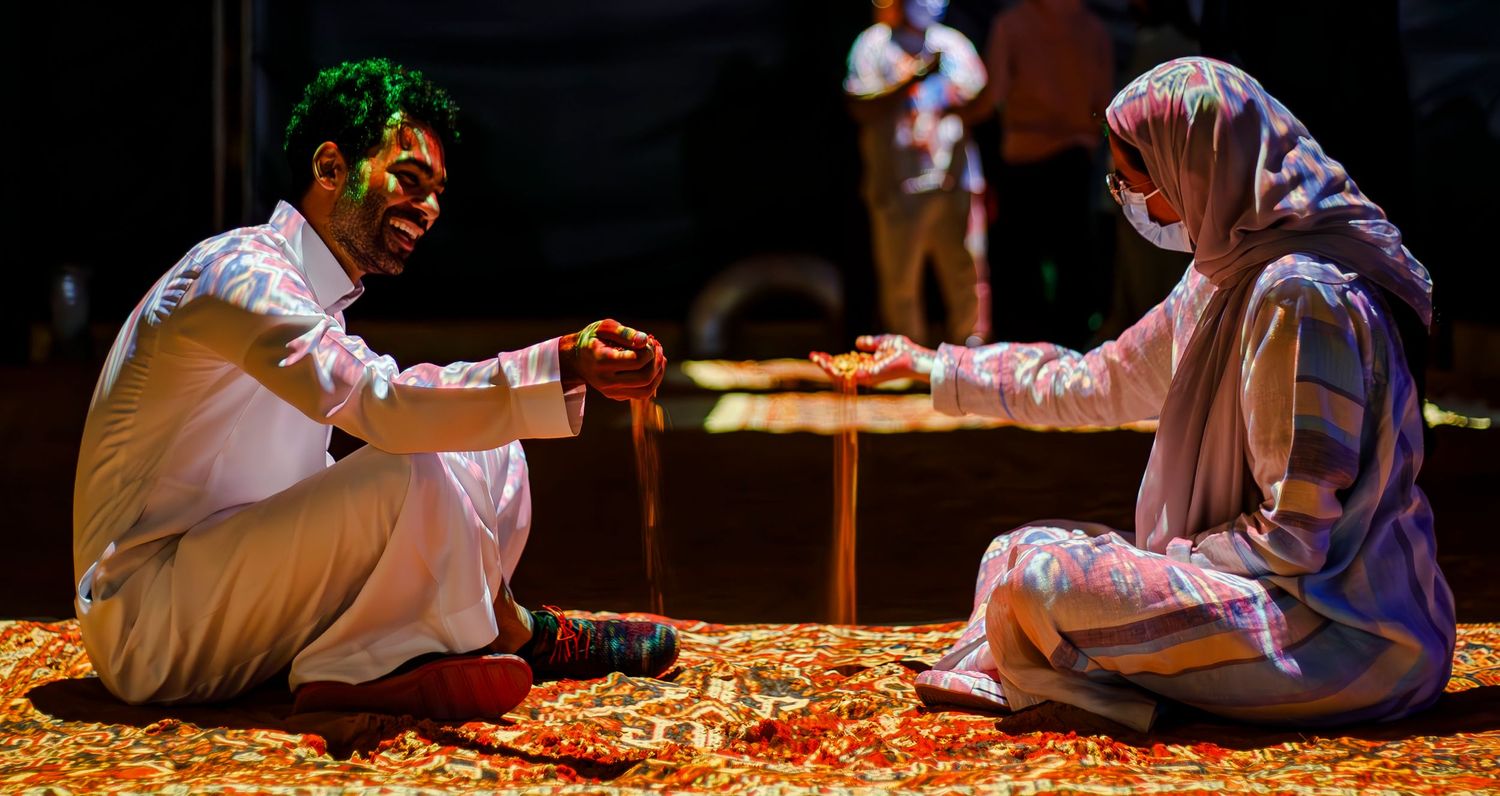 Two people share a traditional meal while seated on an ornate carpet in warm lighting.