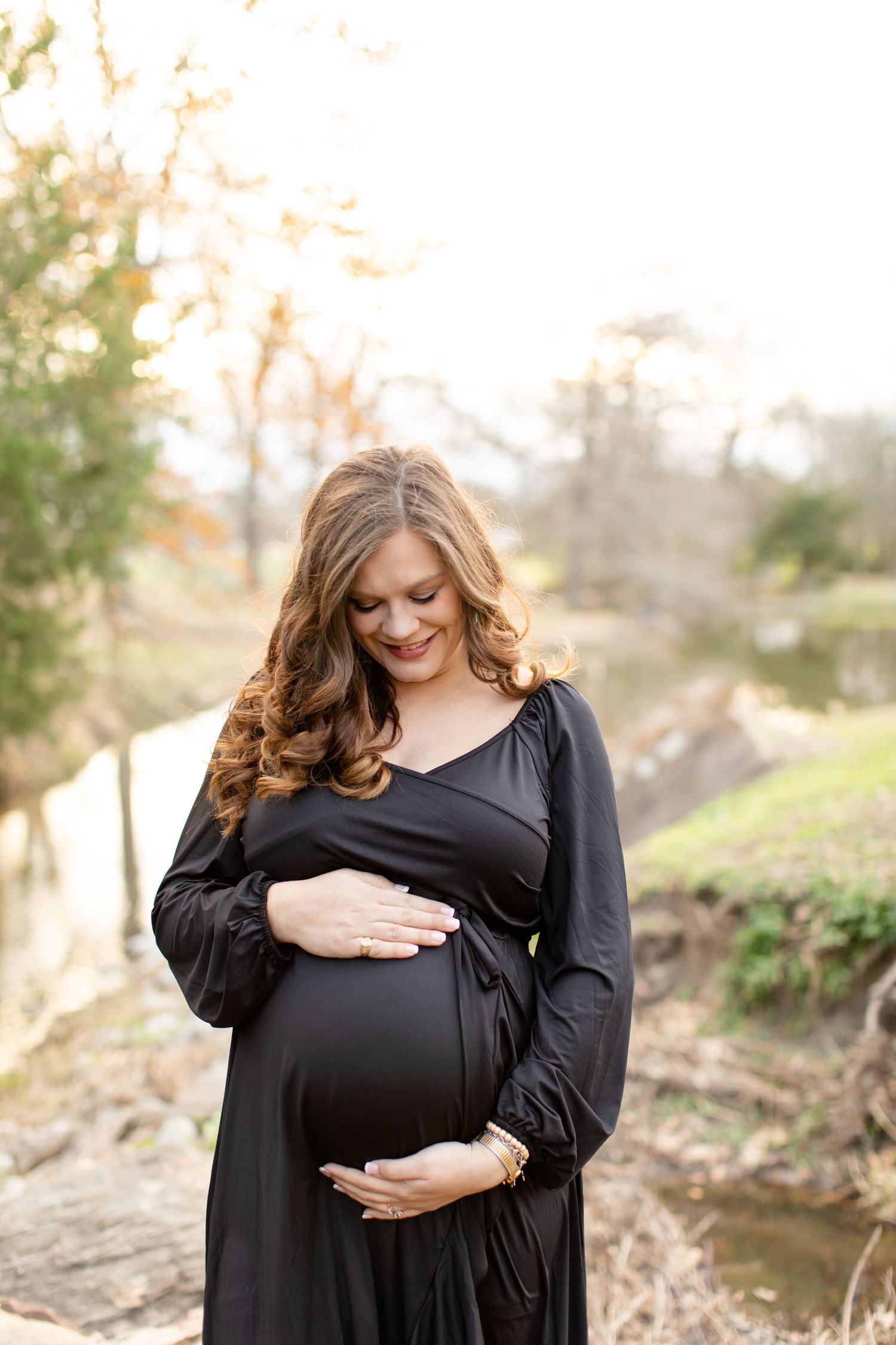 Joyful expectant mother in black dress posing by creek during sunset.
